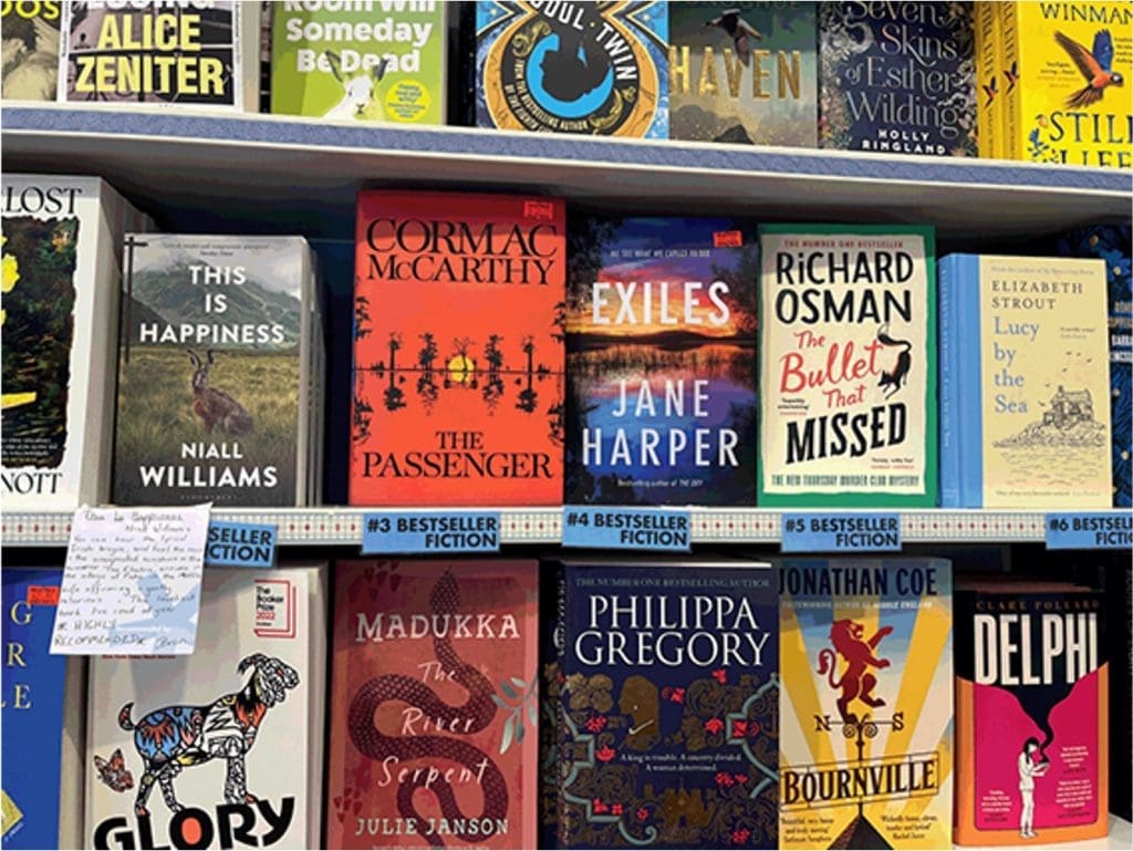 Colorful bookstore shelf with a row of book covers, including The Passenger, Exiles, The Bullet That Missed, and Lucy by the Sea on prominent display under a labeled display rail.
